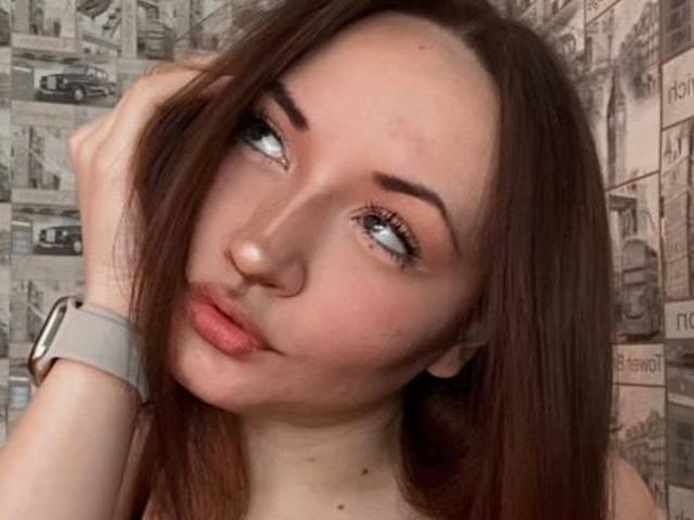 Foto em close-up de RonnieWeeda, uma mulher branca com cabelos castanhos longos, olhos azuis e lábios cheios. Ela está olhando para o lado, com a mão no cabelo, e usando um smartwatch. O fundo tem arte urbana nas ruas. Ela está fazendo um biquinho levemente, transmitindo uma vibe sexy e sedutora..