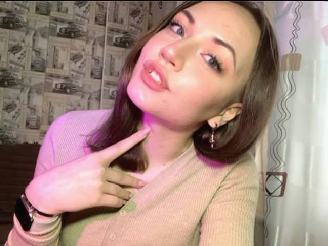 RonnieWeeda hat braunes, schulterlanges Haar, blasse Haut und volle Lippen, die leicht geöffnet sind. Sie trägt einen beigen Pullover, die Hand liegt in der Nähe ihres Halses, und sie blickt verführerisch zur Seite. Der Hintergrund hat ein Zeitungsmuster..