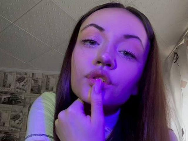 Fotografía de primer plano de RonnieWeeda, una mujer morena con cabello largo, rostro iluminado con luz azul, y un dedo presionado contra su mejilla. Labios entreabiertos, irradiando un aura sensual y provocativa. La uña es morada..