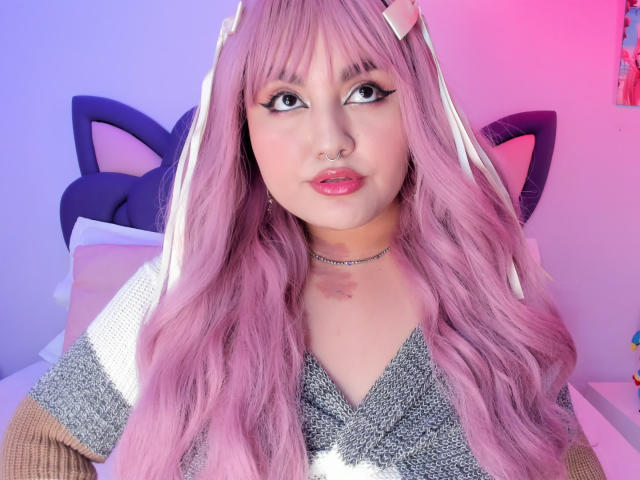 MarlaBriar, avec sa longue perruque rose, ses oreilles de chat et son maquillage prononcé, regarde directement l'appareil photo. Haut argenté scintillant, éclairage rose et bleu, collier ras du cou, tension sexuelle, mignon mais provocateur..