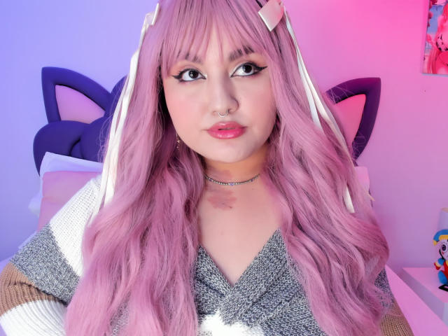 Close-up of MarlaBriar with pink wavy hair, cat ears, and a nose ring. She wears a glittery top, choker, and has a seductive look with pink and blue lighting. Fucking hot.