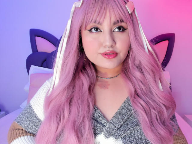 Photograph of MarlaBriar with long, pink, wavy hair, wearing a sparkly gray top and cat ear headband. Pink and purple lighting, sexy expression, subtle makeup, thick black eyeliner.