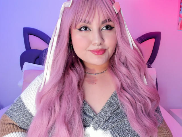 MarlaBriar, met lang roze haar en kattenoren, zit zelfverzekerd, gekleed in een glinsterend grijze top en een choker. Fel roze en blauwe verlichting accentueren haar gezicht, waardoor een sexy en speelse sfeer ontstaat..