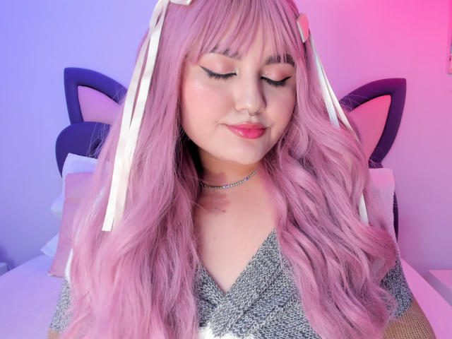 Foto van MarlaBriar met lang roze haar, gesloten ogen en een neusring. Ze draagt een glinsterende top en een choker, en zit tegen een hoofdsteun met kattenoren, terwijl er roze en blauw licht van haar afstraalt. Heel schattig.