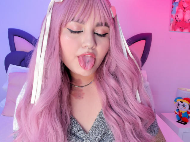 MarlaBriar hat pinkfarbenes, welliges Haar und eine Nasenring, und sie steckt ihre Zunge heraus, die ebenfalls eine Piercing hat. Sie trägt Katzenohren in Schwarz und eine Halskette. Neonrosa und -blaue Beleuchtung. Plüschtier auf der rechten Seite.