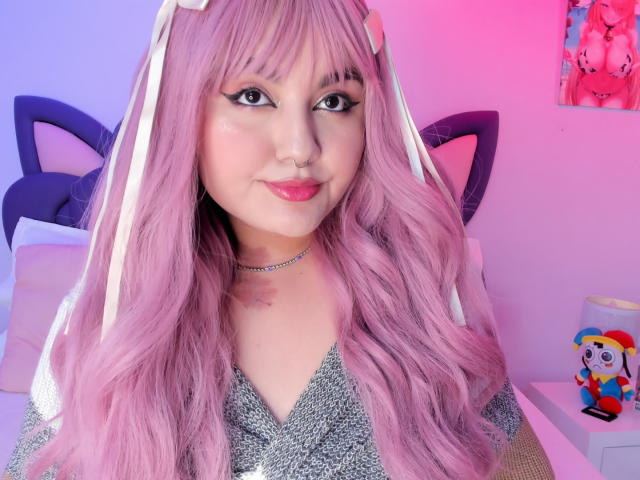 Close-up of MarlaBriar with pink wavy hair, cat ears, and a nose ring, smiling slightly. She wears a glittery top, pink and blue lighting, and a colorful plush toy on the right.