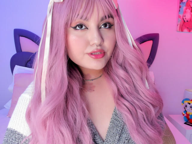 Primo piano di MarlaBriar con lunghi capelli rosa ondulati, che indossa una maglietta grigia scintillante e una collana, e guarda direttamente la fotocamera. Illuminazione rosa e viola, fascia per capelli a forma di orecchie di gatto, labbra leggermente socchiuse..