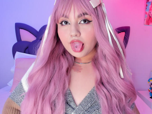 MarlaBriar has long, pink, wavy hair, sticking out her tongue with a silver tongue piercing. She wears a cat ear headband, choker, and glittery top. Neon pink and blue lights in background.