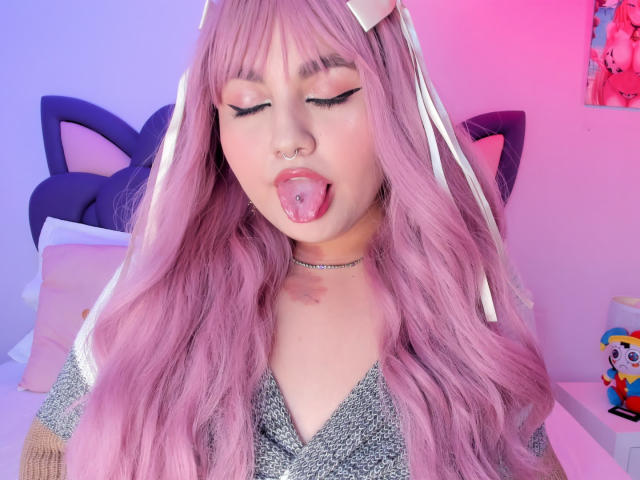 Close-up photo of MarlaBriar, a young woman with pink hair, cat ears, and a nose ring, sticking out her tongue, eyes closed, wearing a sparkly top, neon pink and blue light.