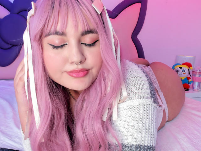 MarlaBriar, uma jovem com cabelo rosa e orelhas de coelho brancas, está deitada em uma cama com os olhos fechados, vestindo uma blusa brilhante. Seu grande bumbum visível, com um brinquedo colorido ao fundo..