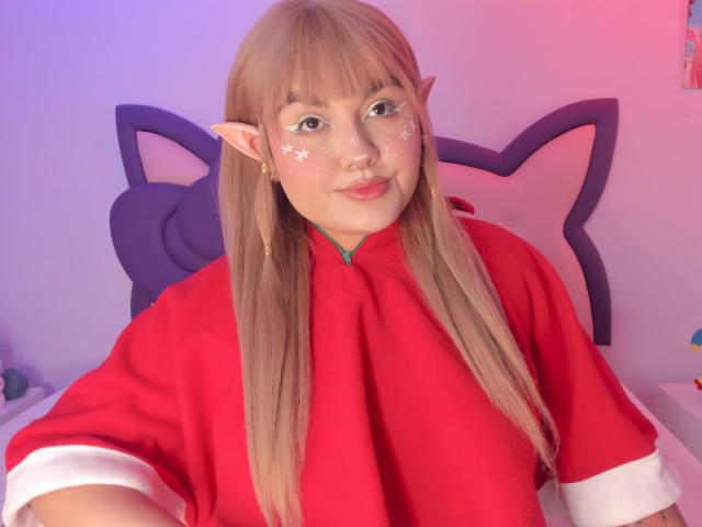 MarlaBriar, com longos cabelos loiros e orelhas de elfo, veste uma roupa de cosplay vermelha com detalhes brancos. Luzes rosa e azul vibrantes realçam seu rosto, marcado com padrões de estrelas brancas. Muito fofo.