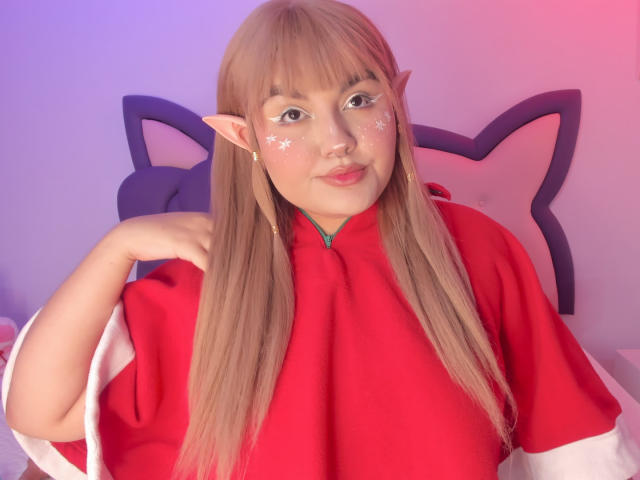 MarlaBriar, con orejas de elfo y cabello rubio largo, lleva un disfraz de cosplay rojo y blanco. Marcas brillantes en el rostro, expresión burlona, mano cerca del cuello. Iluminación en tonos rosados y azules suaves. Muy lindo..