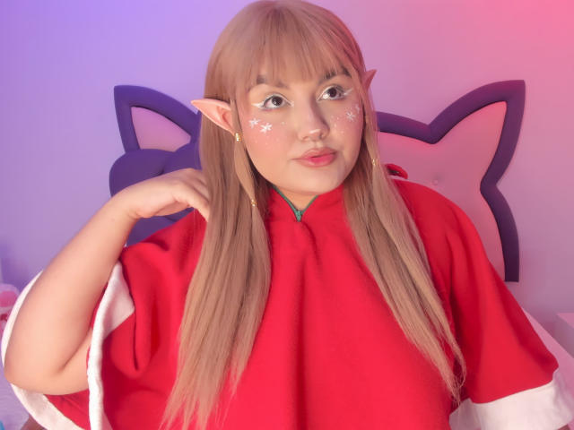 Photograph of MarlaBriar, a chubby woman with long blonde hair, elf ears, and red cosplay outfit. She's posing with a hand near her face, looking up with a slightly flustered expression.