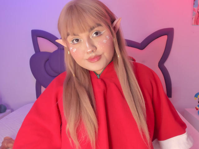 Photographie de MarlaBriar, une elfe cosplay mignonne avec de longs cheveux blonds, portant une cape rouge, des marques faciales en forme d'étoile, et oreilles pointues. Éclairage rose et bleu, pénis visible sur le lit.