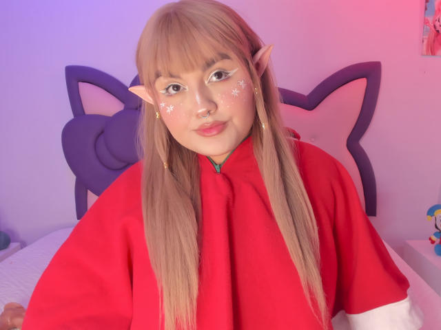 Fotografia di MarlaBriar, un cosplay di una sexy elfa con lunghi capelli castani, una veste rossa, orecchie a punta, stelline adesive sulle guance, e un tocco di rosa. e con luci viola, seduto sul letto con una testiera a forma di gatto..