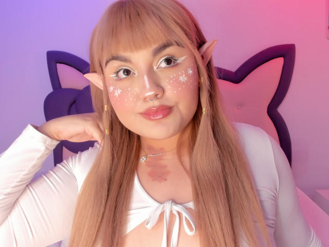 Photograph of MarlaBriar, a young woman with long brown hair, elf ears, and white face glitter, wearing a white shirt, leaning on a pink star-shaped headboard, looking seductive.