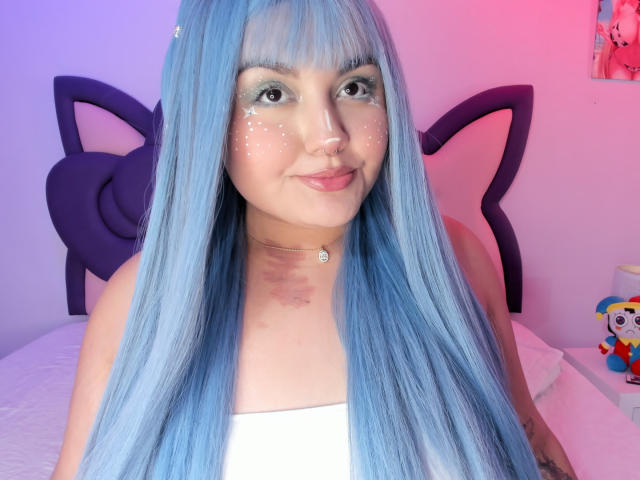 Photograph of MarlaBriar with long, straight, blue hair, wearing a white top, smiling at the camera. Pink and blue light illuminates her face, with a cat-shaped headboard behind her.