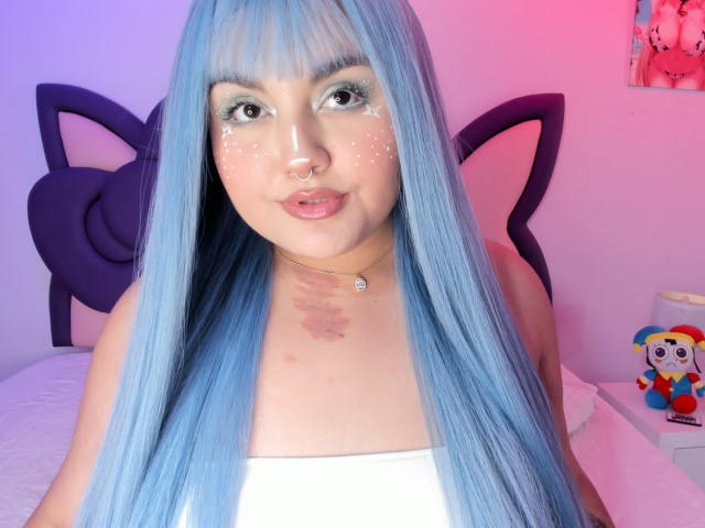 MarlaBriar, avec ses longs cheveux bleus, porte un haut blanc, a un piercing au nez et des paillettes sur ses joues. L'arrière-plan comprend un oreiller en forme de tête de chat noir et un jouet en peluche coloré. Éclairage lumineux.