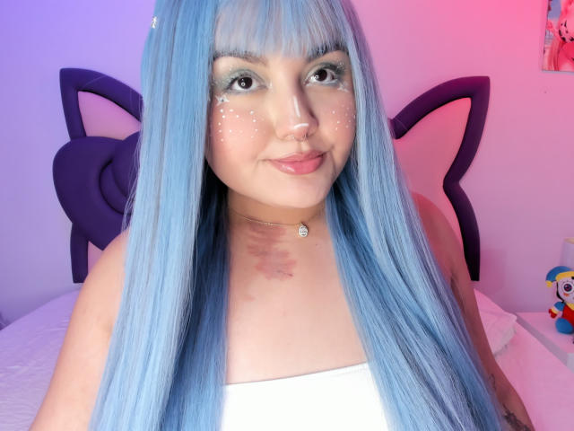 Foto van MarlaBriar met lang, steil, blauw haar, die een witte top draagt, een hoofdtooi met kattenoren en glinsterende make-up. Ze heeft een schattige glimlach, een neusring en een ketting. Er is roze en blauwe verlichting..