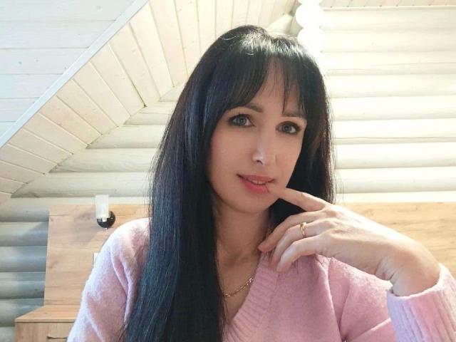 DollishAlisha: atractiva, con cabello negro largo, flequillo y un suéter rosa claro, mordiendo su dedo de forma seductora, ojos marrón claro, rubia. collar, fondo con paneles de madera, luz brillante, ligera sonrisa, pose casual y sensual..