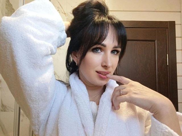 DollishAlisha, bellissima con i capelli neri, avvolta in un asciugamano bianco, con una mano sollevata verso la testa, guarda la fotocamera con un leggero sorriso. Luce soffusa, porta di legno sullo sfondo. Incredibilmente sexy..