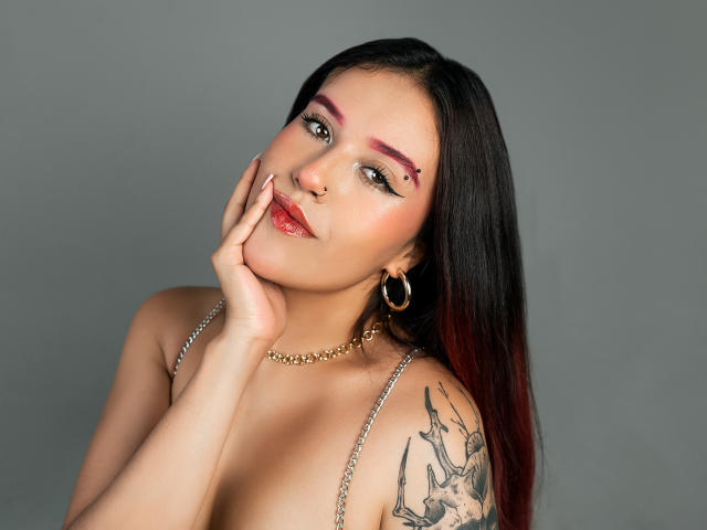 Fotografia di MiaDesiire con lunghi capelli neri, pelle chiara e ombretto rosa. È senza maglietta, con una mano sul viso, indossa una collana a catena d'oro, grandi orecchini a cerchio e ha un tatuaggio floreale sulla spalla. Incredibilmente attraente..