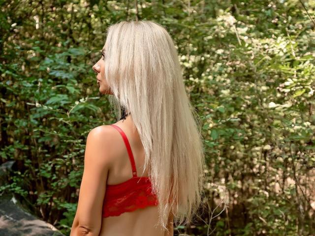 Fotografía de MoniqueBliss, cabello rubio largo, sostén rojo, perfil lateral, fondo de bosque, luz del sol filtrándose a través de los árboles. hojas, sombra en su hombro derecho, ambiente tenso, cabello iluminado por el sol, follaje verde, ambiente veraniego..