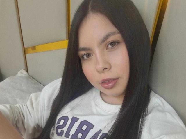 Fotografia de CamiiVasquez com cabelos pretos longos, pele clara e olhos castanhos, vestindo uma camisa branca com a inscrição "LAC". Ela está olhando para a câmera com uma expressão levemente sedutora. O fundo tem painéis amarelos e brancos..