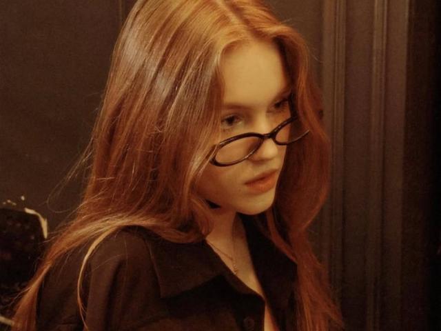Fotografia di Corina Makinster, una giovane donna con lunghi capelli rossi lisci, che indossa occhiali con montatura nera e un abito nero. camicia. Ha un leggero sorriso, e guarda in basso. Illuminazione calda e soffusa. Incredibilmente sexy..