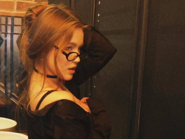 Photographie de Corina Makinster, en profil, séduisante, portant des lunettes noires et un haut noir à une épaule, un collier ras du cou. Cheveux bruns clairs attachés en un chignon désordonné, main derrière la tête, éclairage tamisé, regard séduisant, lumière chaude..