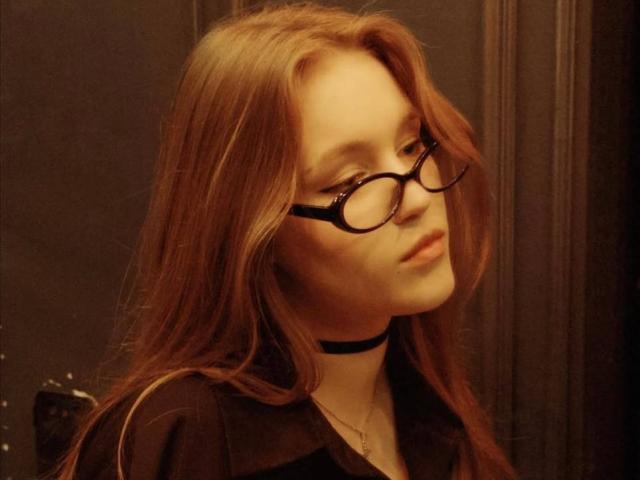 Corina Makinster, met lang, rechtbruin haar, draagt zwarte brillen en een zwarte choker, en kijkt op met een licht verdwaasde blik.
ze draagt een zwarte blouse, die warm gloeit..