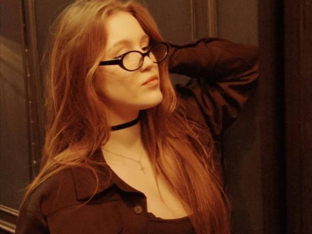 Fotografía de Corina Makinster, una mujer de piel clara con cabello largo y rojo, que lleva gafas negras y un collar negro. Está vestida con una camiseta negra, con la mano detrás de la cabeza, expresión sensual, iluminación tenue..