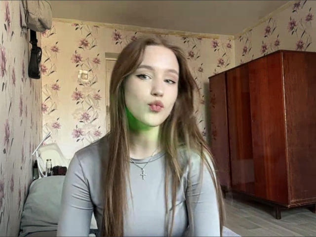 Photograph of CorinaMakinster with long brown hair, wearing a gray long-sleeve top, pouting with slightly pursed lips, standing in a floral-patterned room with wooden furniture. Sexual tension, slight green lighting.