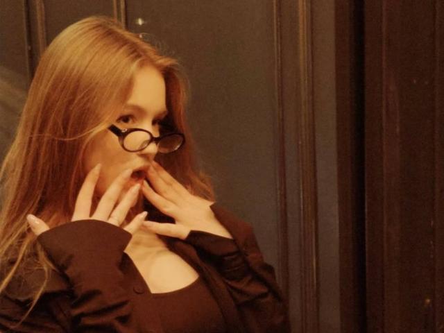 Corina Makinster, mit langem, braunen Haar und einer Brille mit schwarzem Rahmen, bedeckt ihren Mund mit ihren Händen und trägt ein schwarzes Oberteil. Ihr Ausdruck ist scheu, und die Beleuchtung ist warm und gedämpft..