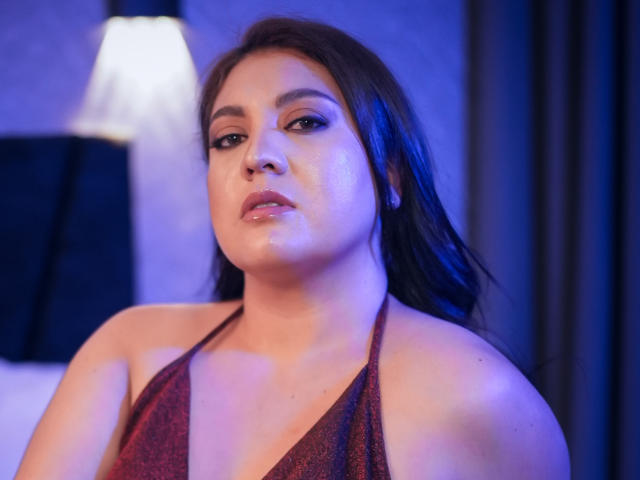 Fotografia em close-up de PaulaAnd, uma mulher de cabelos longos e escuros, vestindo uma blusa vermelha escura com brilhos e alças, olhando sedutivamente para... A câmera capturou um leve tom azulado na pele do rosto dela..