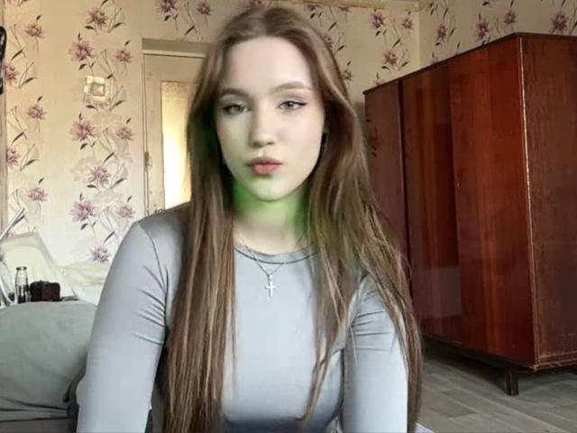 CorinaMakinster with long brown hair, pale skin, and blue eyes, wearing a tight gray top, sitting in a floral-patterned room with wooden furniture. Sexual tension evident in her direct gaze.