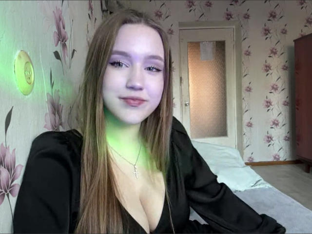 Foto van Corina Makinster, met bleke huid en lang bruin haar, gekleed in een zwarte top met een diepe halslijn, groen licht. verhelderende gezichtsuitdrukking, bloemenbehang, deur op de achtergrond, subtiele glimlach, seksuele spanning, lichte decolleté.