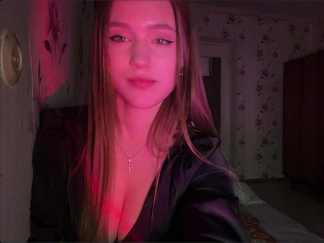 Fotografía de Corina Makinster, mirando a la cámara con iluminación rosa y roja. Cabello largo y castaño, top negro con escote pronunciado, pequeño collar con una cruz. Tensión sexual, expresión ligeramente seductora, habitación con poca luz..