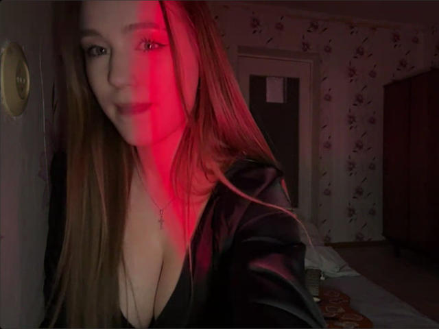 Photograph of CorinaMakinster, illuminated by red light, showing cleavage in a black top, with long brown hair, and a necklace. Dark room, floral wallpaper, door, and bed in background.
