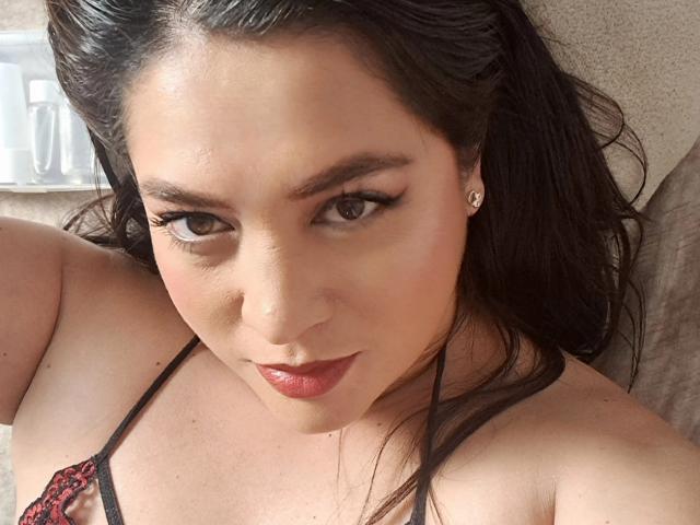 Close-up foto van PaulaAnd, een aantrekkelijke vrouw met donker haar, bruine ogen en rode lippenstift. Ze draagt een zwart en rode kanten BH en kijkt recht in de camera met een verleidelijke uitdrukking..