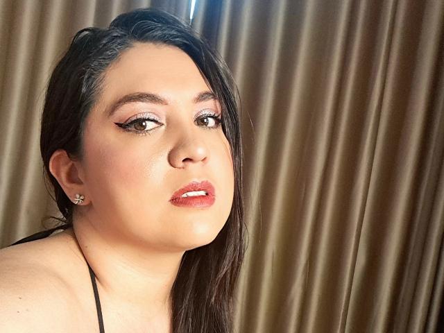 Foto em close-up de Paula, uma mulher latina com cabelos longos e pretos, delineador marcante e batom vermelho, olhando ligeiramente para o lado com uma expressão sugestiva, em frente a cortinas marrons. Muito atraente..