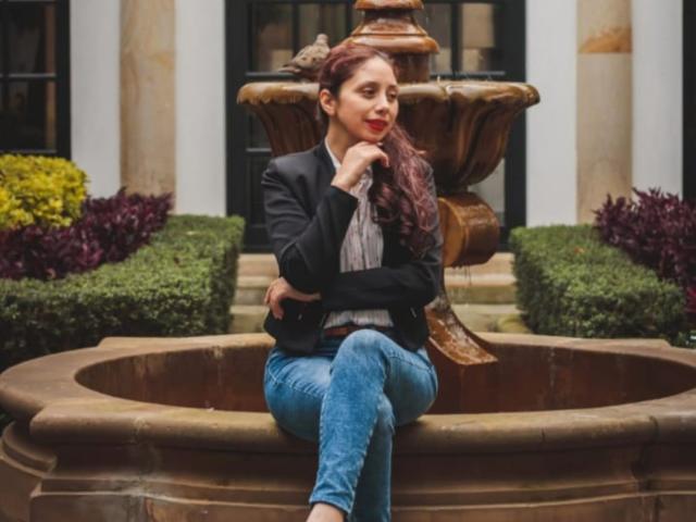 Stephanie Sun, met lang bruin haar, rode lippenstift, een zwart colbert, een wit shirt en blauwe jeans, zittend op een bronzen fontein. hand voor het kin, armen gekruist, verleidelijke blik, buitenomgeving, weelderige begroeiing.