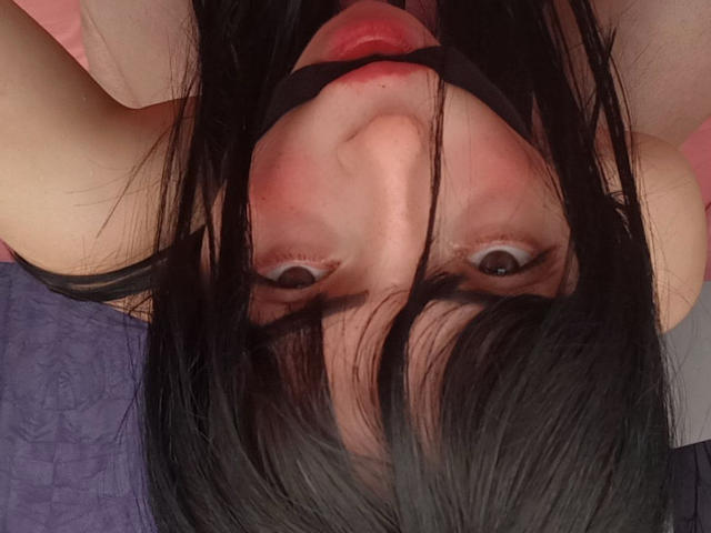 Fotografía de primer plano de Liiil, boca abajo, con el cabello negro cubriendo su rostro, ojos grandes, labios rojos y hombros desnudos. La expresión de Liiil es intensa, lo que crea una sensación de tensión sexual. Borde gris.