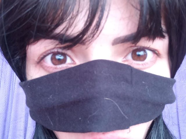 Primo piano di Liiil con capelli neri, occhi marroni e una maschera nera che copre la parte inferiore del viso. La luce intensa evidenzia lo sguardo penetrante di Liiil. Il tessuto texturizzato della maschera è visibile..