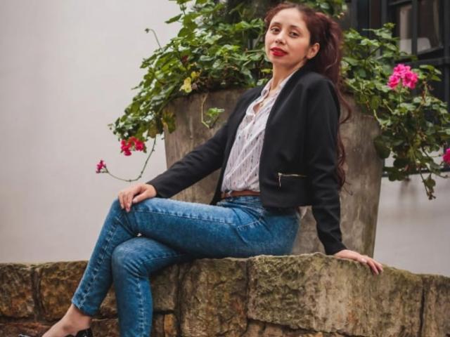 Stephanie Sun, eine braunhaarige Frau mit langen Haaren, sitzt auf einer Steinkante, trägt eine schwarze Jacke, ein weißes Hemd, blaue Jeans und hat lange Beine. gerötete, rote Lippen, Pflanzen mit rosa Blüten im Hintergrund, sexy, selbstbewusste Pose..