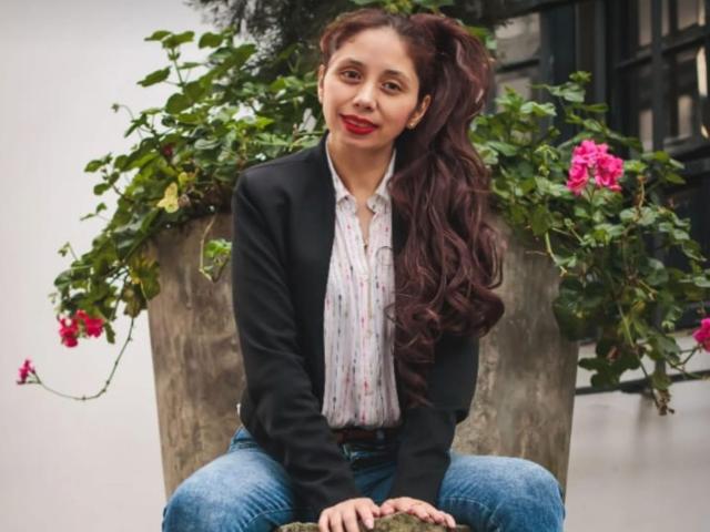 StephanieSun sits with legs spread, wearing a black blazer and white shirt, blue jeans, long dark brown hair in a ponytail, red lips, green leafy plant with pink flowers behind her.