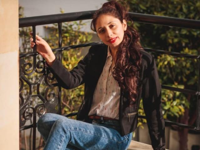 Stephanie Sun, una mujer morena con cabello largo y ondulado, se apoya en una barandilla de hierro negro, vistiendo un blazer negro y una camisa blanca. Una camisa abotonada y pantalones vaqueros azules, con una expresión seductora y segura. La luz del sol resalta su rostro..