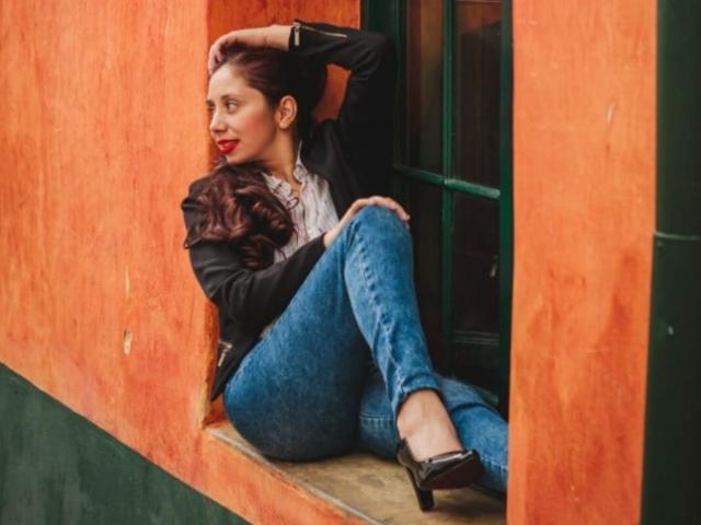 Stephanie Sun, com cabelos castanhos longos, batom vermelho, blazer preto, camisa branca, jeans azuis e saltos pretos, sentada contra um parede laranja, mão na cabeça, pernas cruzadas, olhar intenso, clima sensual..