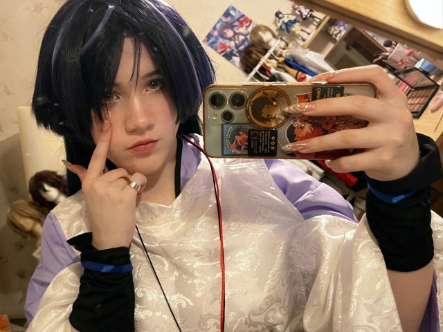 KatlynRenteria, con cabello negro y piel pálida, se toma un selfie frente al espejo, vistiendo una blusa blanca y semitransparente y pantalones negros. pulseras. Su dedo toca su mejilla, mientras sostiene un teléfono con una funda detallada..