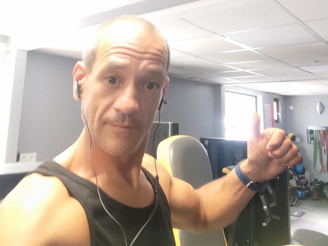 Foto van een kale, middelbare man met een lichte snor, die een zwart tanktop en oordopjes draagt en een vuist omhoog steekt. met een nonchalante grijns. Kantooromgeving.